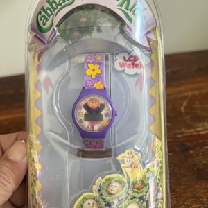 May include: A Cabbage Patch Kids LCD watch in its original packaging. The watch has a purple band decorated with yellow and pink flowers. The watch face features a Cabbage Patch Kid character. The packaging is clear plastic with the Cabbage Patch Kids logo.