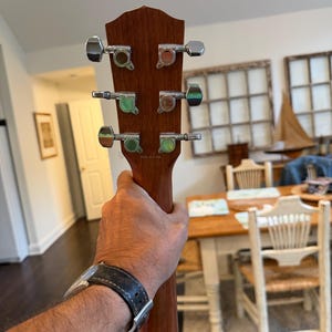 Pu&ograve; includere: Primo piano della paletta di una chitarra acustica marrone. La chitarra ha sei meccaniche cromate con accenti verdi e marroni. La venatura del legno &egrave; visibile sulla paletta e sul manico. La chitarra &egrave; tenuta.