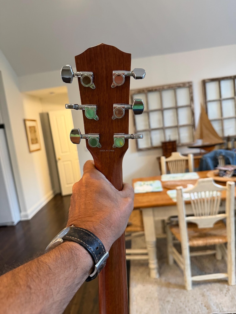 Pu&ograve; includere: Primo piano della paletta di una chitarra acustica marrone. La chitarra ha sei meccaniche cromate con accenti verdi e marroni. La venatura del legno &egrave; visibile sulla paletta e sul manico. La chitarra &egrave; tenuta.