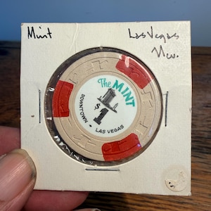 May include: A beige and red casino chip from The Mint in downtown Las Vegas, featuring a black dollar sign and building graphic. The chip is encased in a square cardboard holder with handwritten text reading Mint and Las Vegas Nev.