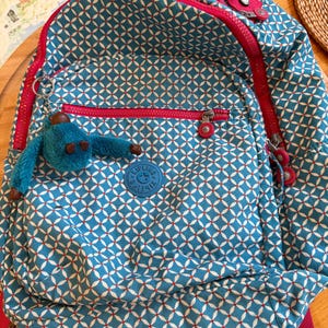 Peut inclure: Sac à dos bleu avec un motif géométrique blanc et rouge. Le sac à dos a une fermeture éclair, des bretelles et une poignée rouges. Un porte-clés singe bleu est suspendu à la poche avant. Le sac à dos a un écusson avec un logo.