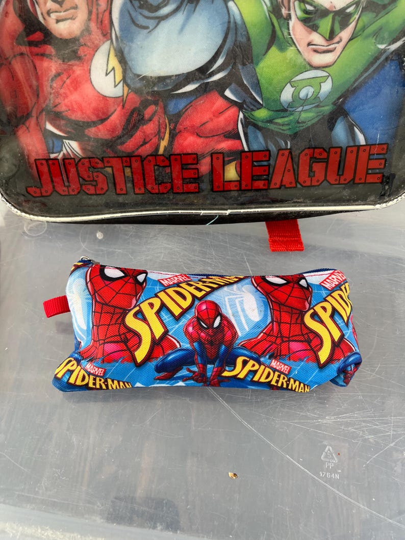 May include: A blue pencil case featuring the Marvel character Spider-Man. The case has a red zipper pull and a repeating pattern of Spider-Man in action poses. The top of the image shows a Justice League lunchbox.