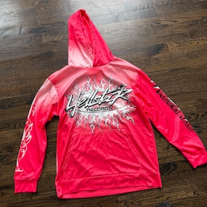 May include: A bright pink long-sleeve hooded sweatshirt featuring a gradient colour effect. The front displays a graphic of a stylised sun with flames and the text Hellstar Records. The sleeves feature additional flame graphics and text.