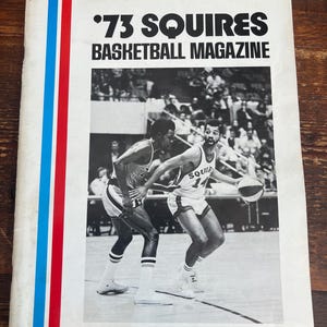 Può includere: Rivista di basket vintage '73 Squires. La copertina presenta uno scatto in bianco e nero di due giocatori di basket in azione. La rivista ha uno sfondo bianco con strisce verticali rosse, bianche e blu sul lato sinistro.