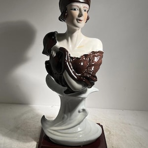 Könnte beinhalten: Keramikbüste einer Frau mit brauner Kopfbedeckung und drapiertem braunem Stoff. Die Büste ist weiß mit braunen Akzenten und steht auf einem dunkelbraunen Sockel. Der Sockel ist rechteckig.