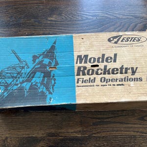 May include: A vintage model rocket kit box. The box is split into blue and tan sections. The blue section features a rocket illustration, while the tan section displays the text "Model Rocketry Field Operations Kit". Recommended for ages 10 to adult.