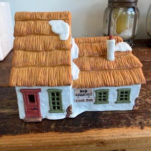 Puede incluir: Figurita de cerámica de una casa de campo con tejado de paja, paredes blancas y marcos de ventanas verdes. La casa tiene una puerta roja y una chimenea. El texto "BOB CRATCHIT TINY TIM" está en el frente. Nieve acentúa el tejado.