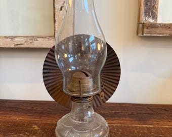 May include: A clear glass oil lamp with a metal base and a clear glass chimney. The lamp has a round, corrugated metal reflector behind the chimney. The lamp sits on a wooden surface. The lamp is a vintage style.