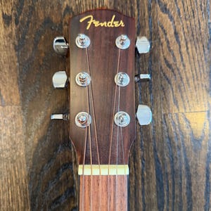 Pu&ograve; includere: Primo piano della paletta di una chitarra acustica Fender. La paletta &egrave; marrone scuro con il logo Fender in oro. Sono visibili sei meccaniche argentate, insieme alle corde e a una parte della tastiera.