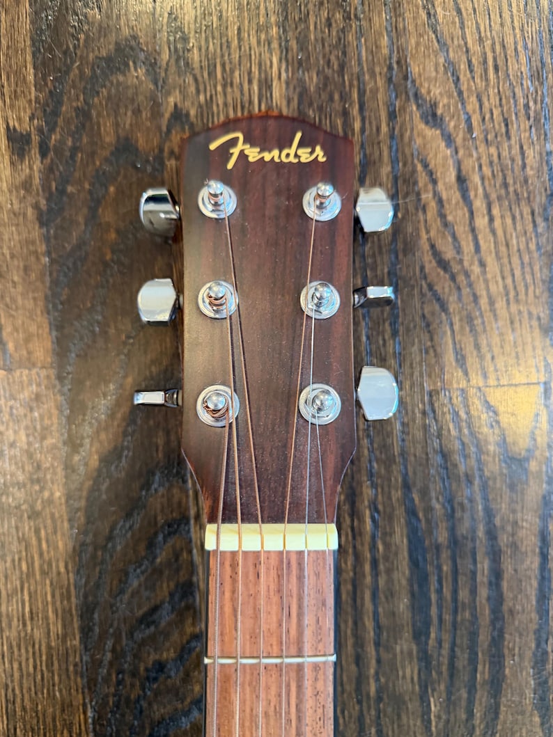 Pu&ograve; includere: Primo piano della paletta di una chitarra acustica Fender. La paletta &egrave; marrone scuro con il logo Fender in oro. Sono visibili sei meccaniche argentate, insieme alle corde e a una parte della tastiera.