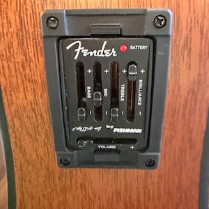 Pu&ograve; includere: Primo piano di un pannello di controllo del preamplificatore per chitarra Fender Classic 4 by Fishman nero. Presenta cursori per bassi, medi, alti e brillantezza, insieme a un controllo del volume e un indicatore della batteria. Il pannello &egrave; montato su un corpo di chitarra in legno.