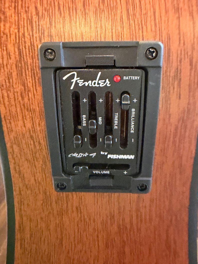 Pu&ograve; includere: Primo piano di un pannello di controllo del preamplificatore per chitarra Fender Classic 4 by Fishman nero. Presenta cursori per bassi, medi, alti e brillantezza, insieme a un controllo del volume e un indicatore della batteria. Il pannello &egrave; montato su un corpo di chitarra in legno.
