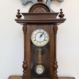 May include: A brown wooden wall clock featuring a circular white face with black Roman numerals and ornate black hands. The clock has a glass-fronted lower section displaying a gold-toned pendulum with the letters R and A inscribed on it.