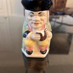 Puede incluir: Figura de cerámica de un hombre con vestimenta tradicional, que incluye un sombrero negro y un abrigo verde. La figura sostiene una pipa. La figura tiene una cara pintada y está sentada.