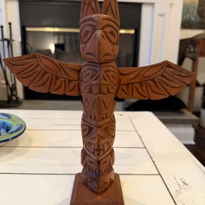 May include: A carved wooden totem pole with a reddish-brown finish. The totem features a bird-like figure at the top, outstretched wings, and carved faces. The base is a rectangular block. The background includes a fireplace and a white table.
