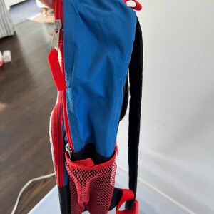 May include: A small, colorful backpack with a blue main compartment, red accents, and a black back and straps. Features a red mesh side pocket and a zippered front. The zipper has a red pull tab.