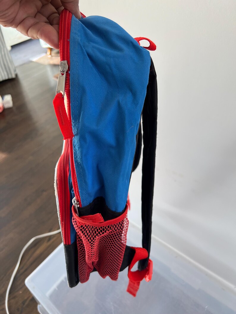 May include: A small, colorful backpack with a blue main compartment, red accents, and a black back and straps. Features a red mesh side pocket and a zippered front. The zipper has a red pull tab.
