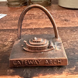 Puede incluir: Un organizador de escritorio de metal color bronce con forma de Gateway Arch. La base es rectangular con las palabras "GATEWAY ARCH" en relieve. El arco se eleva desde la base, con una representación detallada de un edificio.