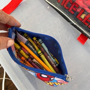 May include: A blue pencil case with a Spider-Man design, filled with crayons and a pencil. The case has a zipper closure and a red strap. The case is open, revealing the colorful contents.