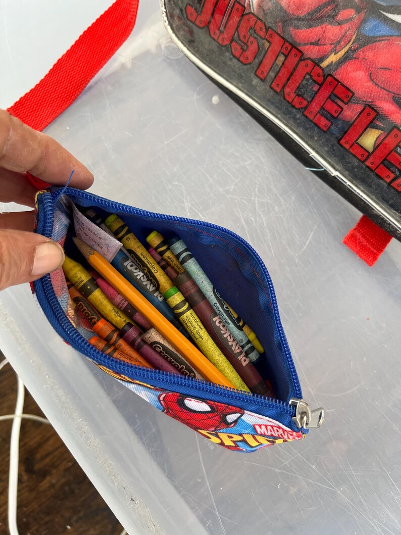 May include: A blue pencil case with a Spider-Man design, filled with crayons and a pencil. The case has a zipper closure and a red strap. The case is open, revealing the colorful contents.