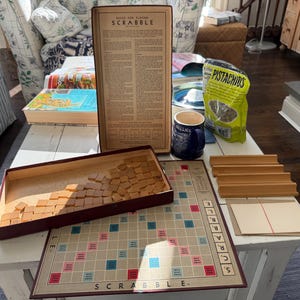 May include: A vintage Scrabble board game set with a maroon box of letter tiles, a game board, wooden tile racks, and a rule book. A blue mug and a bag of pistachios are also on the table.