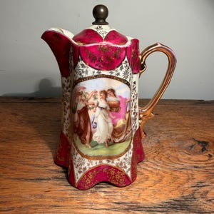 May include: A porcelain coffee pot featuring a deep magenta and gold floral pattern with a central oval medallion depicting three figures in classical attire. The pot has a scalloped base, a gold-toned handle, and a dark brown spherical lid finial.