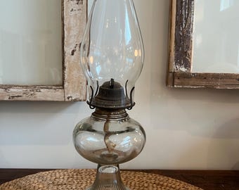 May include: A clear glass oil lamp with a bulbous base and a tall, flared chimney. The lamp has a metal burner and a wick visible inside. The base is ribbed, and the lamp sits on a woven mat. The background includes a window frame.