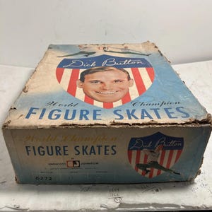 Może przedstawiać: Zabytkowe pudełko na łyżwy figurowe z portretem Dicka Buttona, mistrza świata w łyżwiarstwie figurowym. Pudełko jest niebiesko-białe z czerwonymi akcentami, a tekst "World Champion Figure Skates" jest widoczny.