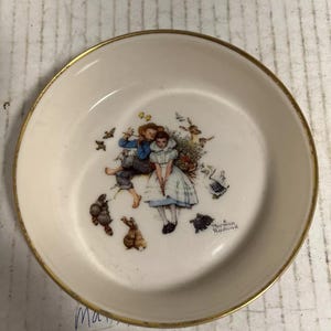 May include: A small, round, off-white dish with a gold rim. The centre features a colourful illustration of children and animals. The artwork includes a boy and girl, rabbits, and birds. The dish is likely vintage.