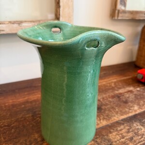 May include: A green ceramic vase with a glossy finish, featuring a wide, flared, and wavy rim. The upper section of the vase includes two small, circular holes integrated into the rim structure, providing a distinct opening for decorative use.