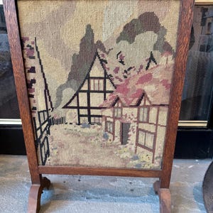 May include: A wooden fire screen with a tapestry showing a landscape scene with houses and trees. The screen has a rectangular frame and two small feet. The tapestry features shades of beige, brown, and pink.