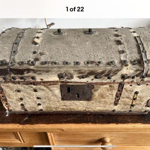 May include: An antique trunk with a weathered, textured exterior. The trunk is rectangular with a curved top, reinforced with metal bands and rivets. A dark metal lock is centered on the front. The trunk sits on a wooden surface.