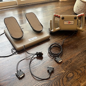 May include: A beige Pro Pedals foot pedal set with two pedals and a matching steering wheel controller. The controller has red buttons and the text "Virtual Pilot II". Black cables and connectors are attached.