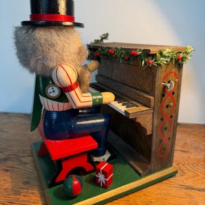 May include: A wooden nutcracker figure wearing a black top hat and green vest, seated on a red bench playing a brown upright piano. The piano is decorated with a green garland and red berries. A small red gift box and a ball sit on the green base.