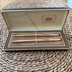 Könnte beinhalten: Ein goldfarbenes Stift- und Bleistiftset in einer schwarz-beigen Box. Die Box ist geöffnet und zeigt die Schreibgeräte auf einer braunen, samtartigen Oberfläche. Auf der Box steht das Wort "CROSS".