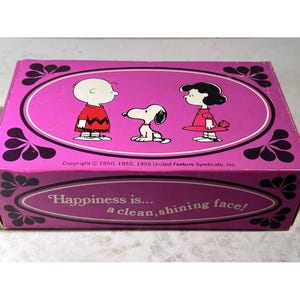 Pode incluir: Caixa rosa com os personagens Peanuts, Charlie Brown, Snoopy e Lucy. A caixa tem um design oval com o texto "Happiness is... a clean, shining face!" e informações de direitos autorais.