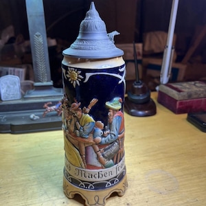 Puede incluir: Un jarro de cerveza vintage con tapa de peltre y una escena en relieve detallada de personas bebiendo. El jarro tiene un fondo azul oscuro con un diseño de sol dorado. La base tiene las palabras "Machen froh".