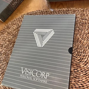 以下が含まれることがあります： 白い幾何学模様のVisiCorp Personal Softwareの灰色の箱。箱には水平線とVisiCorpのロゴがあります。他のソフトウェアボックスが背景に見えます。