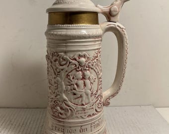 Puede incluir: Jarra de cerveza de cerámica color crema con un diseño en relieve detallado y una tapa con bisagras. La jarra presenta un asa y un relieve decorativo con figuras y texto. La tapa tiene una banda dorada.