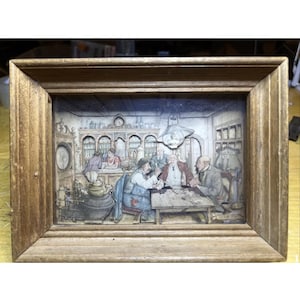 May include: A framed, three-dimensional artwork depicting a detailed interior scene with figures. The artwork is encased in a wooden frame. The scene features people in a room with tables, shelves, and various objects, creating a historical or vintage aesthetic.