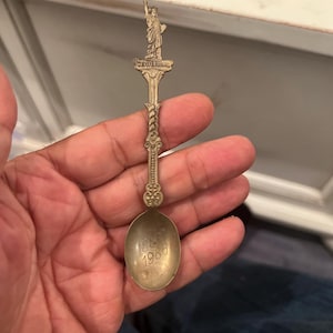 May include: A vintage souvenir spoon with a detailed Statue of Liberty figure at the top. The spoon is made of a gold-colored metal and features the engraved dates 1886 and 1980 on the bowl. The spoon is held in a hand.