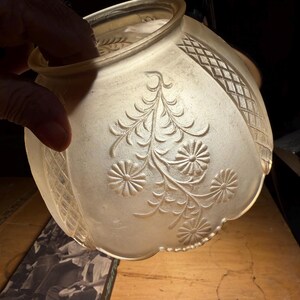 May include: Frosted glass lampshade with a floral and geometric design. The shade has a scalloped edge and is held by a hand. The floral design includes detailed flowers and leaves.