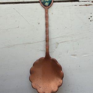 May include: A vintage copper spoon with a decorative handle and a scalloped bowl. The handle features a small shield-shaped emblem with the word "Colorado" and a landscape scene. The spoon is likely a souvenir or collectible item.