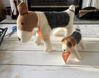 Può includere: Due statuette di cani in feltro. Il cane più grande ha un corpo bianco, la schiena nera e la testa marrone. Il cane più piccolo ha una combinazione di colori simile. Entrambi i cani hanno etichette arancioni. I cani sono su una superficie bianca.