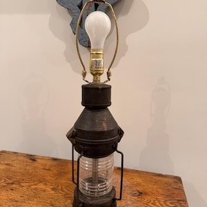 Op de afbeelding: Een lamp in antieke stijl met een heldere glazen cilinder, ingesloten in een donker metalen frame. De lamp heeft een messingkleurige lamphouder en een witte gloeilamp. De lamp staat op een houten oppervlak.