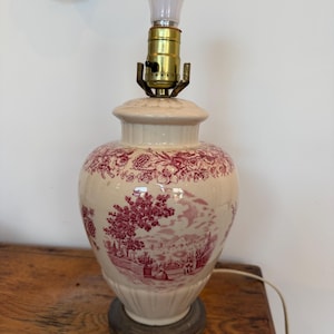 Puede incluir: Lámpara de mesa de cerámica color crema con un diseño floral y paisajístico rojo. La lámpara tiene una bombilla, un accesorio de latón y una base decorativa. La lámpara es de estilo vintage y es adecuada para una sala de estar o dormitorio.