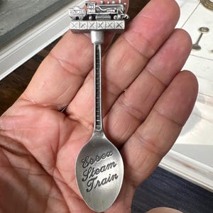 Puede incluir: Cuchara de recuerdo plateada con una locomotora en miniatura detallada sobre un puente. La cuchara lleva grabada la inscripción "Essex Steam Train" en escritura decorativa, un objeto de colección.