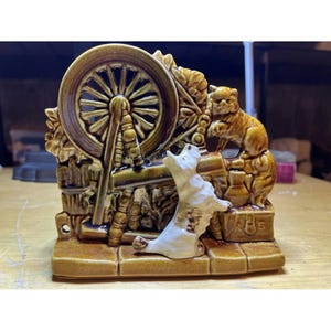 May include: A ceramic figurine depicting a spinning wheel with a dog and a cat. The figurine is primarily brown and white, with intricate details. The dog is brown and the cat is white.