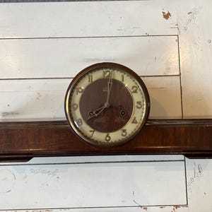 May include: A vintage wooden mantel clock with a round face and a dark brown finish. The clock face has a light-colored background with black numerals and hands. The clock sits on a rectangular base.