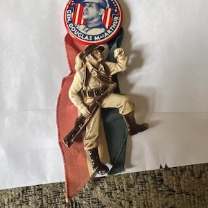 Op de afbeelding: Een vintage General Douglas MacArthur-speld. De speld heeft een cirkelvormig ontwerp met het portret en de tekst van MacArthur. Een beeldje van een soldaat met een geweer is aan de speld bevestigd. De speld is bevestigd aan rode, witte en groene linten.